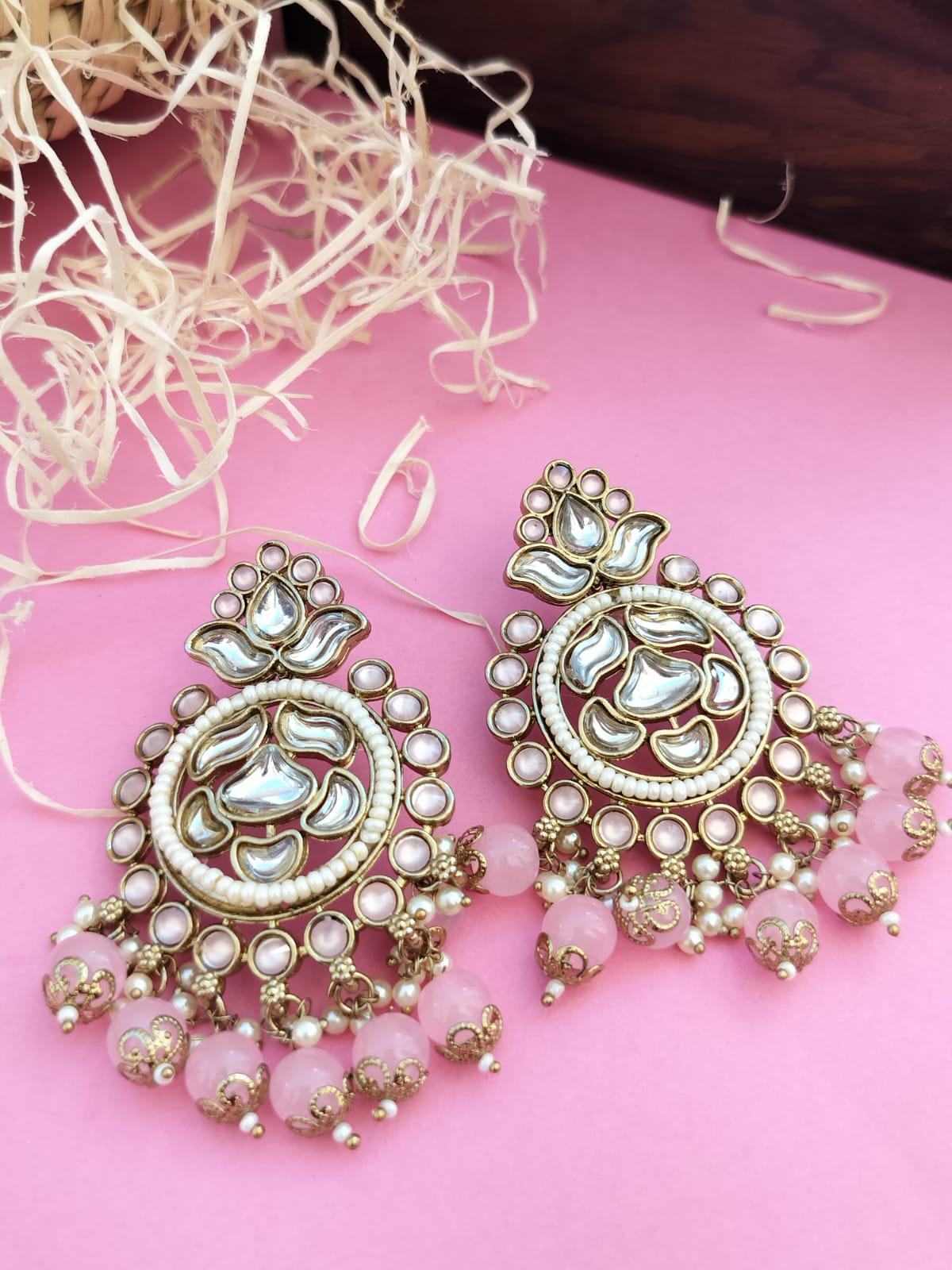 A pair of light pink, gold-colored Kundan Pearl Chandbali earrings with intricate floral patterns and pearl embellishments, displayed on a pink surface with strawberry decorations around.