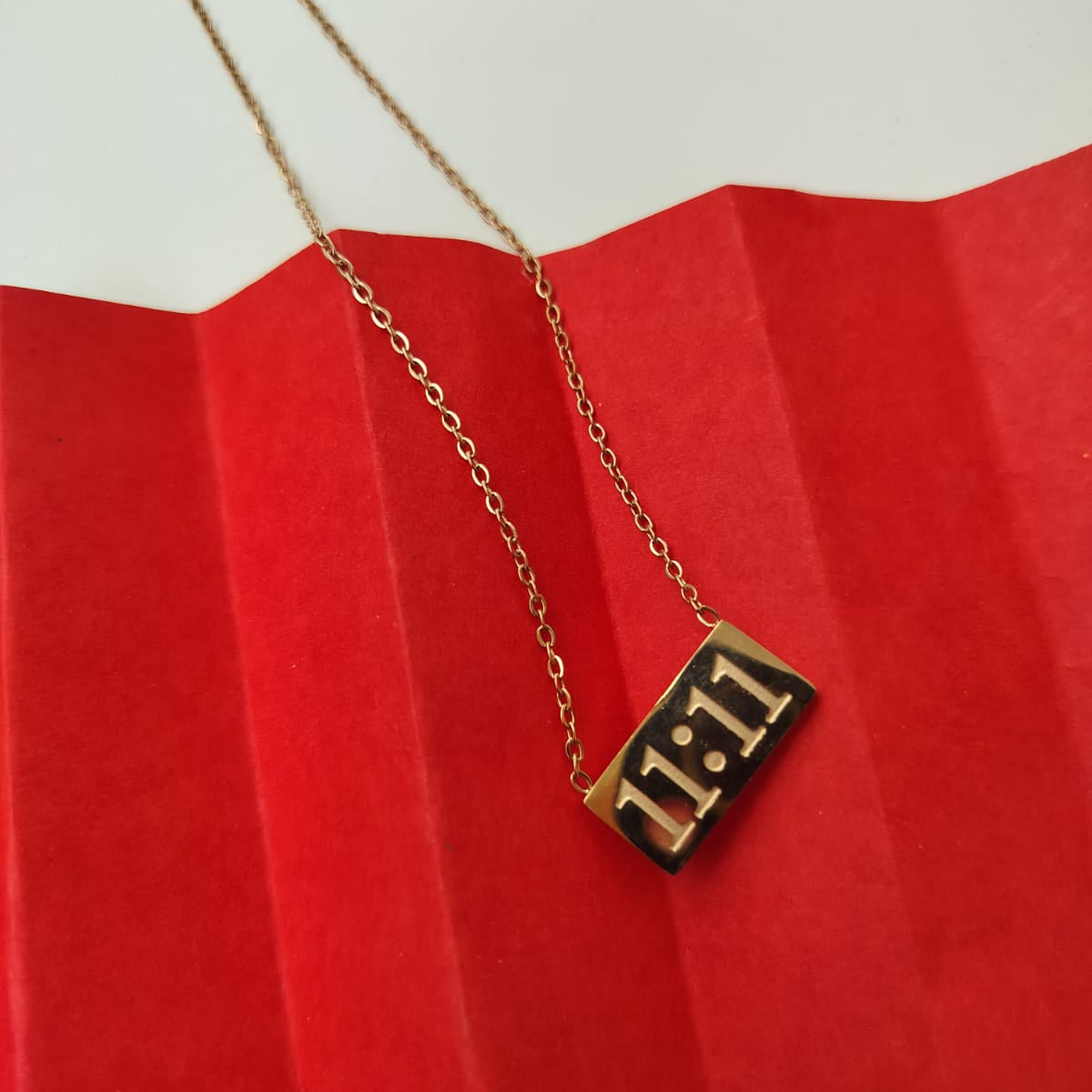 A rose gold pendant with the number 11:11 on it, displayed on a red textured surface, attached to a chain.