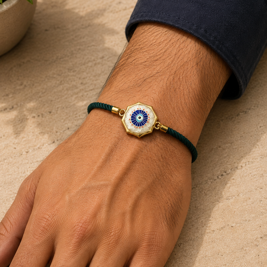 A gold-plated hexagonal charm bracelet with a blue and white evil eye design, worn on a person's wrist with a dark green cord.