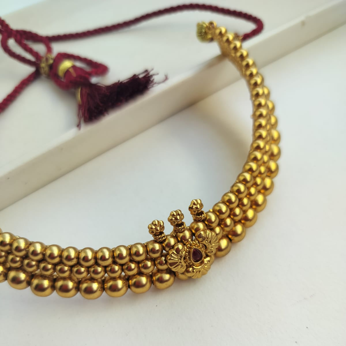A gold-plated beaded necklace with a central decorative filigree piece and a small red stone accent, displayed on a white surface with a maroon tassel cord visible in the background.