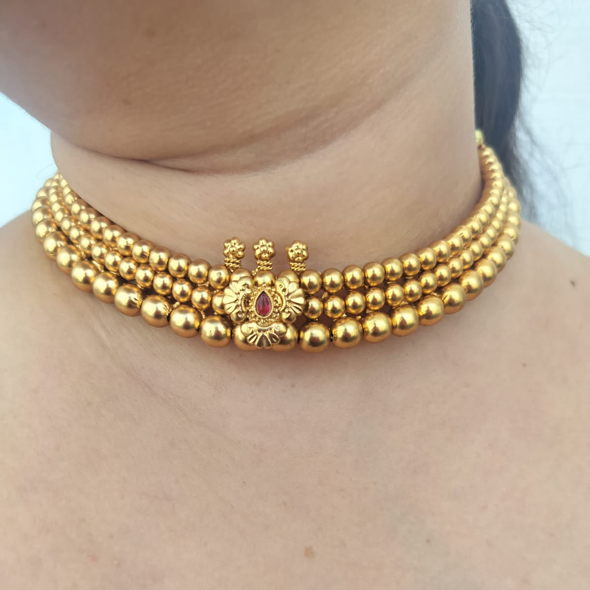 A close-up of a model wearing a gold multi-strand beaded choker necklace with a central decorative gold motif and a small red stone accent.