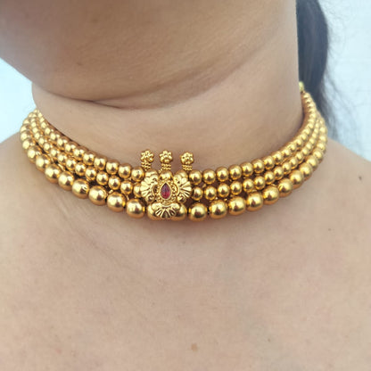 A close-up of a model wearing a gold multi-strand beaded choker necklace with a central decorative gold motif and a small red stone accent.