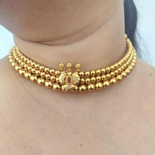 A close-up of a model wearing a gold multi-strand beaded choker necklace with a central decorative gold motif and a small red stone accent.