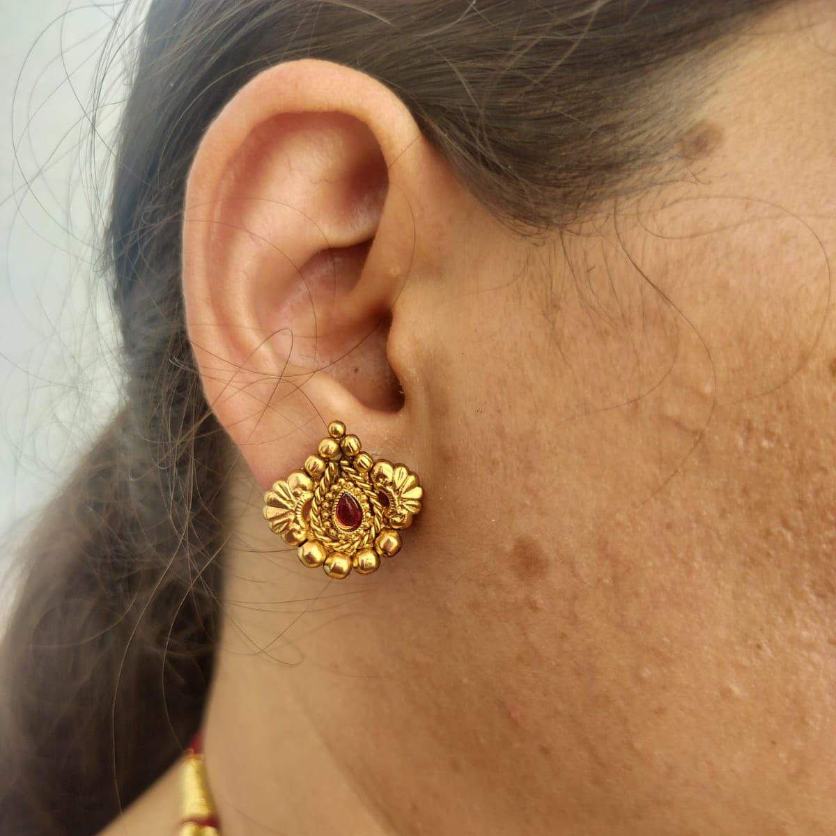 A close-up of a woman's ear wearing a gold traditional stud earring with a small red stone, alongside a matching gold beaded necklace.